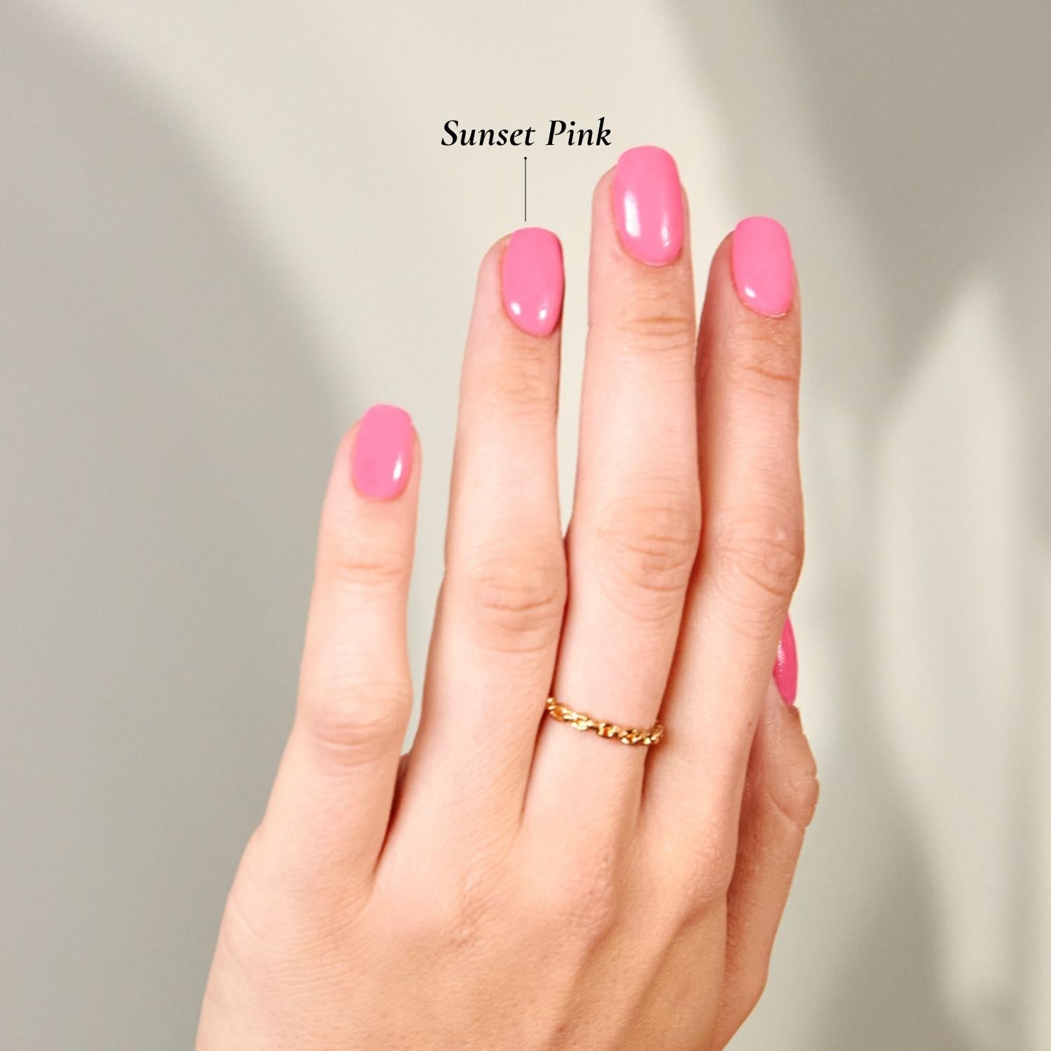 Schlichte, elegante Maniküre in der Farbe 'Sunset Pink' von DIPDRIP. Die oval geformten Nägel erstrahlen in einem glänzenden, kräftigen Pinkton, während ein zarter Goldring das minimalistische Design unterstreicht. Der Hintergrund ist schlicht und lässt die Farbe im Fokus stehen.